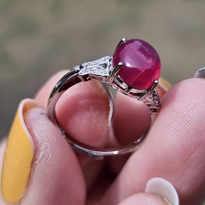 May include: A silver-toned ring featuring a large, smooth, cabochon-cut deep pink gemstone set in a prong setting. The band is accented with small, clear, sparkling stones embedded along the shoulders of the metal frame on both sides.
