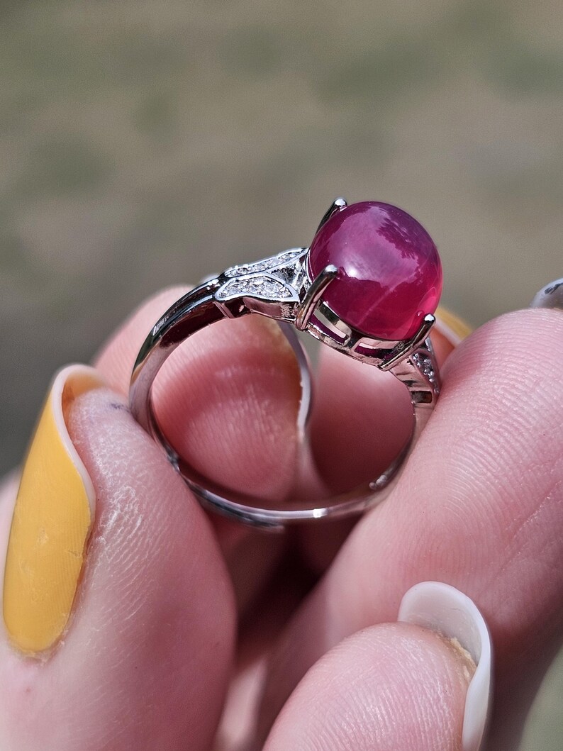 May include: A silver-toned ring featuring a large, smooth, cabochon-cut deep pink gemstone set in a prong setting. The band is accented with small, clear, sparkling stones embedded along the shoulders of the metal frame on both sides.