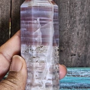 May include: A polished, columnar crystal with a pointed top. The stone features bands of white, gray, and reddish-brown hues. The crystal is held in a hand, showcasing its size and texture. The background is a wooden surface.