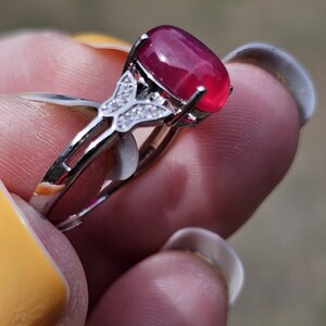 May include: A silver-toned ring featuring a large, oval-shaped, deep pink cabochon gemstone set in a prong setting. The band includes a decorative butterfly-shaped accent on each side, which is embellished with small, clear, sparkling stones.