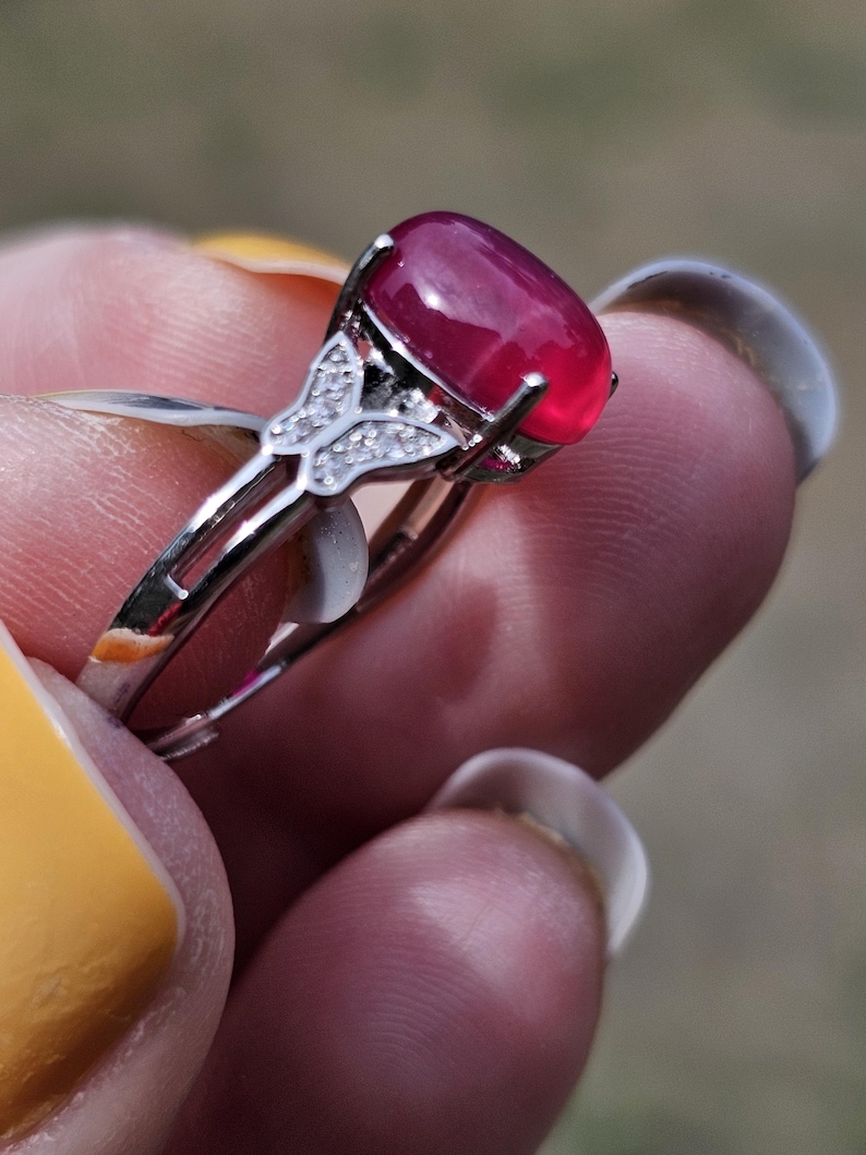 May include: A silver-toned ring featuring a large, oval-shaped, deep pink cabochon gemstone set in a prong setting. The band includes a decorative butterfly-shaped accent on each side, which is embellished with small, clear, sparkling stones.