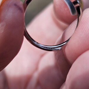May include: A silver-toned adjustable ring featuring a prominent deep red gemstone set in a metal frame. The band is open at the bottom, allowing for size adjustments, and the metal surface has a polished, reflective finish throughout the piece.