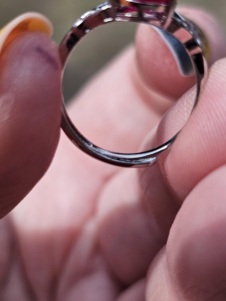May include: A silver-toned adjustable ring featuring a prominent deep red gemstone set in a metal frame. The band is open at the bottom, allowing for size adjustments, and the metal surface has a polished, reflective finish throughout the piece.