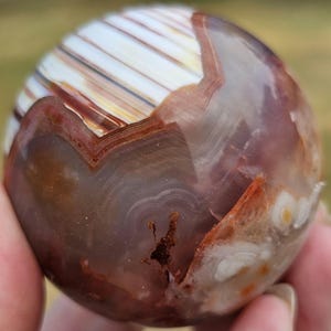 May include: A polished spherical stone featuring a smooth surface with translucent purple and brown tones. The sphere displays distinct white banded layers, intricate swirling patterns, and small dark inclusions within its mineral structure.