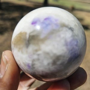 May include: A polished, spherical stone with a marbled appearance. The orb features a base of white and gray hues, accented with patches of purple and brown. The smooth surface reflects light, creating a subtle sheen. The stone is held in a hand.