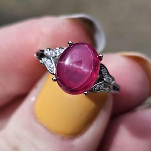 May include: A silver-toned ring featuring a large, oval-shaped, deep pink cabochon gemstone at the center. The band is accented with small, clear, sparkling stones set along the shoulders, creating a delicate, symmetrical leaf-like design.