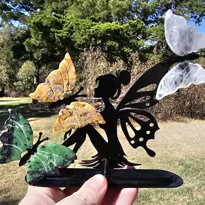 May include: A black metal silhouette of a fairy with wings, featuring three attached butterfly-shaped stones. One butterfly is green with black speckles, two are yellow with brown patterns, and one is translucent white, all mounted on a base.