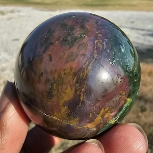 May include: A polished gemstone sphere with a mix of colors including green, purple, yellow, and orange. The sphere is held in a hand and has a smooth, reflective surface. The sphere is a decorative item.