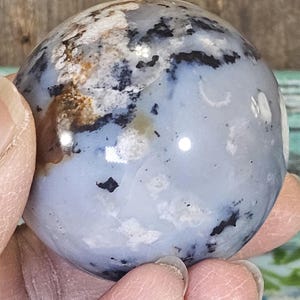 May include: A polished, spherical stone with a mix of colors. The stone features a base of pale blue with swirling patterns of white, black, and brown. The surface is smooth and reflective, with a glossy finish.