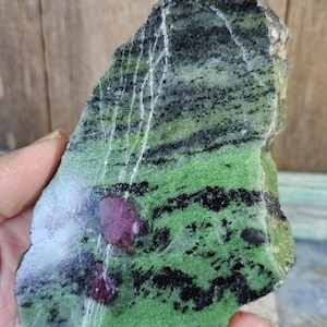 May include: A rough-cut stone featuring a vibrant green base with black and white veining. The stone has patches of deep red ruby crystals. The natural stone is held in a hand, showcasing its unique texture and color variations.
