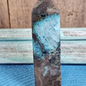 May include: A rectangular crystal obelisk with a pointed top. The obelisk features a striking combination of colors, including shades of brown, teal, and turquoise. The crystal has a textured surface with visible patterns and variations in color.