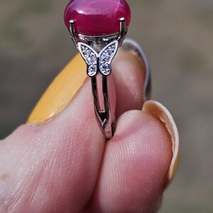 May include: A silver-toned ring featuring a large, smooth, oval-shaped deep pink cabochon gemstone set in a prong setting. The band is accented with a small butterfly motif encrusted with tiny clear stones on each side of the central stone.