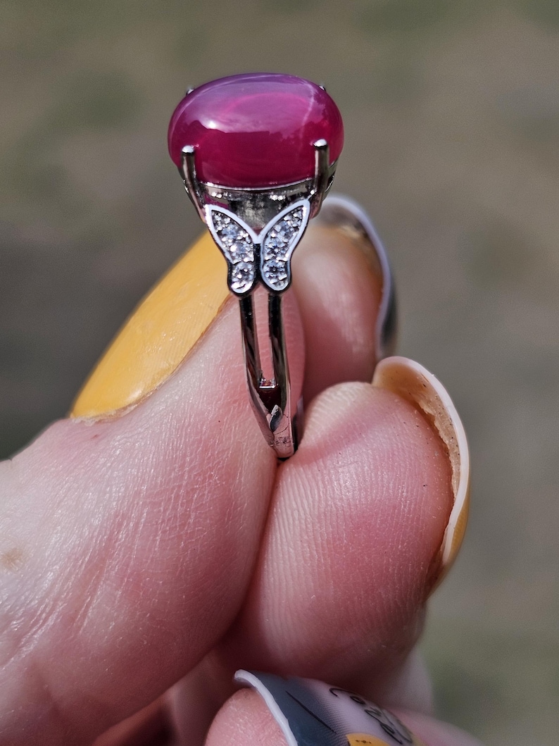 May include: A silver-toned ring featuring a large, smooth, oval-shaped deep pink cabochon gemstone set in a prong setting. The band is accented with a small butterfly motif encrusted with tiny clear stones on each side of the central stone.