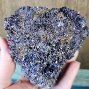 May include: A close-up of a dark blue mineral specimen with a textured surface. The mineral has small, sparkling white and gold flecks throughout. The specimen is held in a hand, showcasing its natural, rough form.