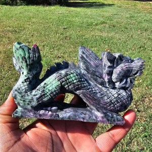 May include: A carved stone dragon figurine in shades of green, purple, and black. The dragon is in a coiled pose, with detailed scales and wings. The figurine is held in a hand against a backdrop of green grass.