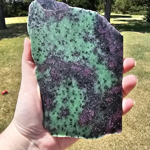 May include: A flat, irregularly shaped slab of zoisite mineral featuring a mottled pattern of vibrant green and deep purple-red inclusions. The surface displays a polished texture with dark, speckled spots distributed throughout the stone.