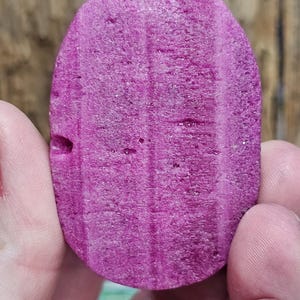 May include: A close-up of a vibrant, oval-shaped, magenta-colored stone. The stone has a textured surface with vertical lines and small holes. The stone is held between two hands.