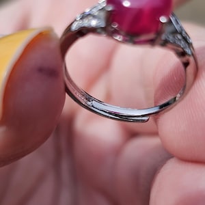 May include: A silver-toned ring featuring a large, smooth, oval-shaped deep pink cabochon gemstone set in a prong setting. The band is adjustable with an open gap at the bottom and includes small, clear, sparkling stones along the shoulders.