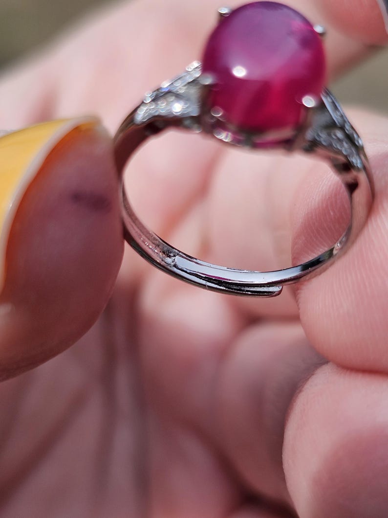 May include: A silver-toned ring featuring a large, smooth, oval-shaped deep pink cabochon gemstone set in a prong setting. The band is adjustable with an open gap at the bottom and includes small, clear, sparkling stones along the shoulders.