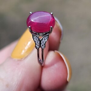 May include: A silver-toned ring featuring a large, smooth, oval-shaped magenta gemstone set in a four-prong mount. The band is accented with a butterfly-shaped design on each side, which is embellished with small, clear, sparkling stones.