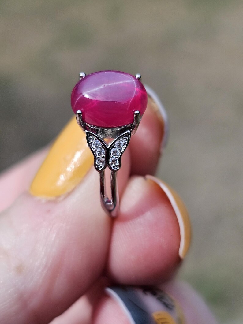 May include: A silver-toned ring featuring a large, smooth, oval-shaped magenta gemstone set in a four-prong mount. The band is accented with a butterfly-shaped design on each side, which is embellished with small, clear, sparkling stones.