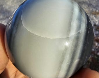 May include: A polished spherical stone featuring smooth, flowing bands of light gray, charcoal, and white. The surface has a reflective, glossy finish that highlights the natural layered patterns and subtle color gradients within the mineral sphere.