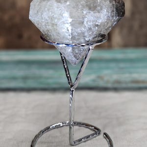 May include: A clear, faceted crystal sits atop a silver metal stand. The stand has a triangular support and a decorative, coiled base. The crystal is a light gray color and has a diamond shape.