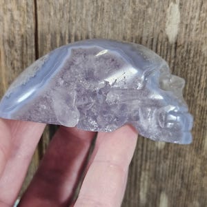 May include: A carved skull figurine made of a translucent, light blue and lavender-colored stone. The skull's features are detailed, with visible eye sockets, a nose, and teeth. The stone has a polished, smooth surface.