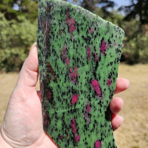 May include: A hand holding a polished slab of zoisite with ruby inclusions. The stone features a vibrant green base color speckled with black and irregular patches of deep pink ruby.