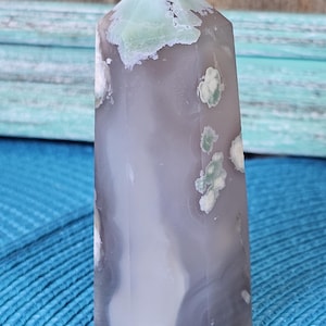 May include: A polished agate crystal point. The crystal features a light gray base with swirling patterns and patches of pale green and white. The top is a light green color. The crystal is displayed on a blue surface.