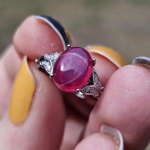 May include: A silver-toned ring featuring a large, oval-shaped, cabochon-cut deep pink gemstone at the center. The band is accented with small, clear, sparkling stones set along the shoulders, creating a decorative, leaf-like metal design.