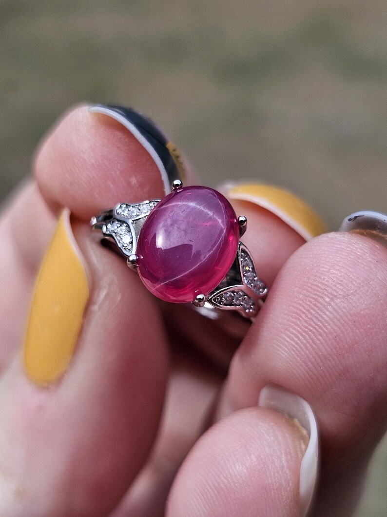 May include: A silver-toned ring featuring a large, oval-shaped, cabochon-cut deep pink gemstone at the center. The band is accented with small, clear, sparkling stones set along the shoulders, creating a decorative, leaf-like metal design.