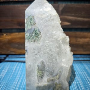 May include: A clear quartz crystal point with green moss-like inclusions. The crystal is cut into a rectangular shape with a pointed top. The crystal is displayed against a blue and brown background.
