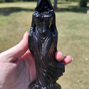 May include: A black figurine depicting a hooded figure in a long, flowing gown. The sculpture features a smooth, dark finish with detailed folds in the fabric of the garment and a deep, shadowed hood covering the upper portion of the figure.