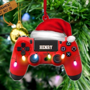 May include: Red and black video game controller Christmas ornament with a Santa hat on top. The controller has a name "HENRY" on it and is decorated with colorful lights.