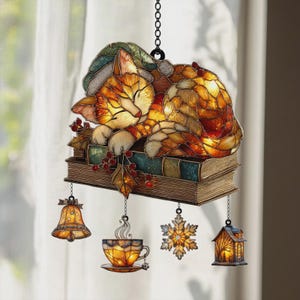 Pode incluir: Enfeite de gato em vitral dormindo sobre livros, com um sino, uma xícara de chá, um floco de neve e uma casa pendurados embaixo. O gato é laranja e branco, usando um chapéu verde. Os livros são verde-azulado e marrom.