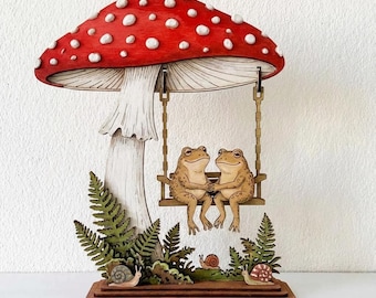 Peut inclure: Objet d'art en bois sur le thème des champignons, avec deux grenouilles sur une balançoire. Le champignon a un chapeau rouge à pois blancs. Des fougères et des escargots sont à la base.