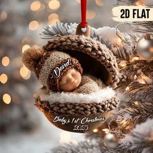 May include: A Christmas ornament featuring a sleeping baby in a knitted acorn-shaped cradle. The baby wears a brown and beige hat with the name "David". The ornament is inscribed with "Baby's 1st Christmas 2025".