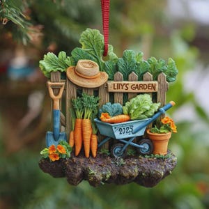 Puede incluir: Adorno con temática de jardín que presenta una escena de jardín en miniatura. Incluye un sombrero de paja, una pala, zanahorias, una carretilla con verduras y una maceta. El adorno tiene el texto "LIIYS GARDEN" y el año "2023".