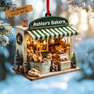 May include: A miniature bakery ornament with a green and white striped awning, labeled "Ashley's Bakery." The ornament features a display of baked goods, small Christmas trees, and a sign that reads "This ornament is flat, not 3D."