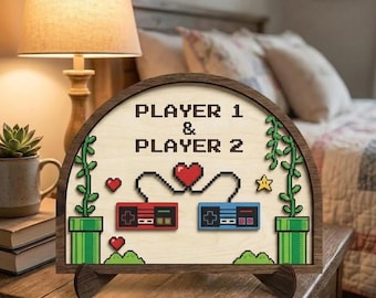 Op de afbeelding: Houten bord met een halfronde bovenkant, met de tekst "PLAYER 1 & PLAYER 2". Het heeft gepixelde gamecontrollers, harten en een ster. Het bord wordt ondersteund door een houten standaard en staat op een houten oppervlak.