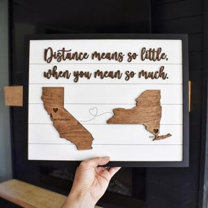 Op de afbeelding: Houten wandkunst met een zwart frame. De kunst toont houten uitsparingen van Californië en New York, verbonden door een stippellijn. De tekst "Distance means so little when you mean so much" staat boven de staten.