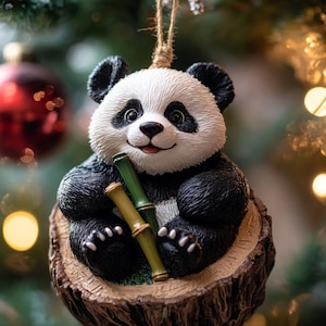 Op de afbeelding: Een hangend ornament met een zwart-witte pandabeer die op een gestructureerde bruine boomstronk zit. De panda heeft een glimlachende uitdrukking en houdt twee groene bamboestengels vast. Het ornament hangt aan een dun, lichtbruin touwtje.