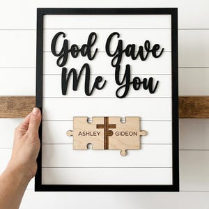 May include: A framed wooden sign with the words "God Gave Me You" in black script. Below, two wooden puzzle pieces with the names "Ashley" and "Gideon" and a cross are displayed. The background is white with horizontal wooden slats.