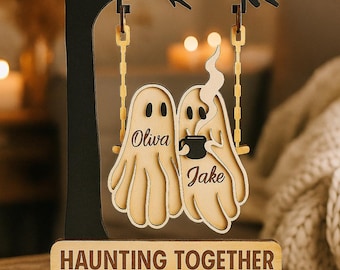 May include: Wooden Halloween decoration featuring two ghost figures on a swing, with the names "Oliva" and "Jake". One ghost holds a coffee cup. The base reads "HAUNTING TOGETHER SINCE 2015". The tree and swing are black and the ghosts are light brown.