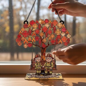 Puede incluir: Un adorno colgante decorativo con un diseño de árbol que presenta emblemas de bomberos y llamas. Las ramas del árbol sostienen bocas de incendio y distintivos de bomberos. Debajo, una pareja con uniformes de bombero está sentada en un columpio. El texto dice "My Hero or Burning Love."