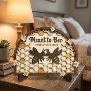 Könnte beinhalten: Holzschild mit dem Text "Meant to Bee" und einem Wabenmuster. Das Schild zeigt zwei Bienen-Silhouetten und ist mit kleinen Gänseblümchen verziert. Das Schild wird auf einem Holzständer präsentiert.