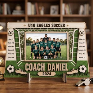 May include: A wooden picture frame shaped like a soccer goal, featuring the text U10 EAGLES SOCCER, COACH DANIEL, and 2024. The frame displays a team photo and lists player names with numbers. A gold MVP trophy and a silver whistle are nearby.
