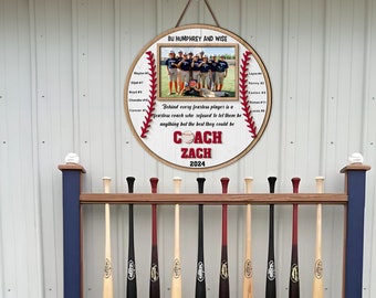 May include: A wooden baseball-themed sign with a team photo, the text "COACH ZACH 2024," and player names. The sign is round with a baseball stitch design. Below, a wooden rack holds several baseball bats.