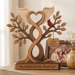 Könnte beinhalten: Holz-Gedenkbaumskulptur mit herzförmigem Design und Engelsflügeln. Ein roter Kardinal sitzt auf einem Ast. Die Basis lautet "Forever In Our Hearts". Ein herzförmiges Etikett lautet "Always With Us 1976-2026."